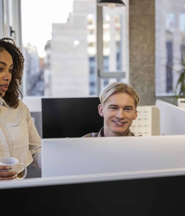 Female customer service representative wearing a headset is holding a mug and assisting a male colleague with a computer issue in a modern office with large windows and natural light
